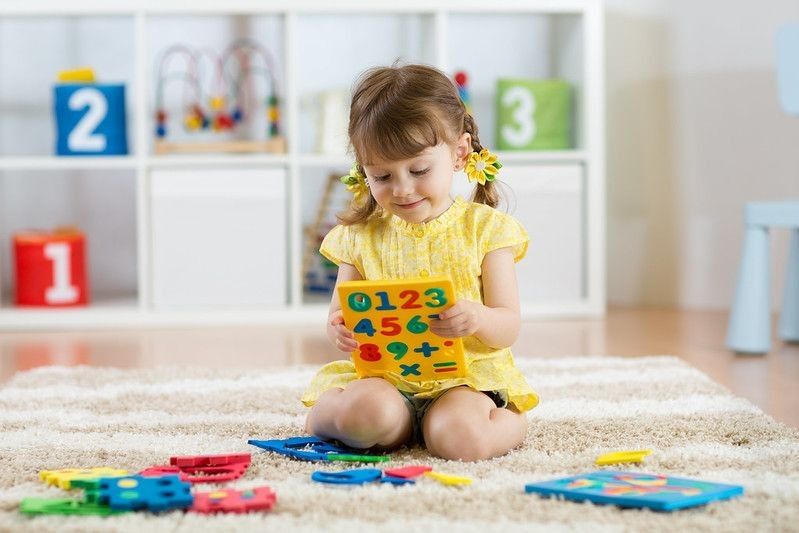 Little girl child playing with lots of colorful plastic digits or numbers on floor indoors.