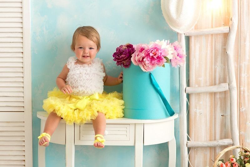 Cute baby girl wearing yellow tulle dress sitting next to flower vase on a table