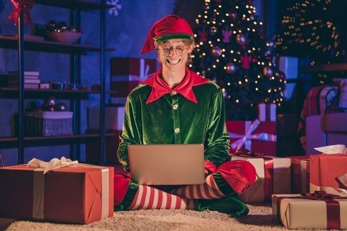 photo of elf sit floor type greeting on laptop wear green glasses cap in house indoors with evening x-mas decor lights