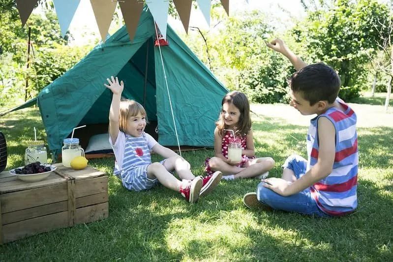 Three kids sat on the grass in front of their camping tent having fun and eating snacks.