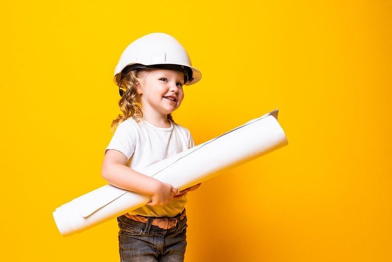 Happy young girl engineer with helmet holding bluepring isolated on yellow background.