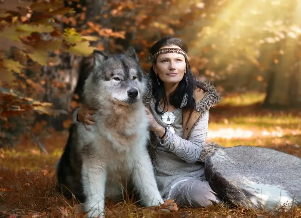 A calm fluffy white and black wolf dog sitting besides a woman in forest
