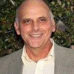 Kurt Fuller standing in front of a leafy background, smiling in a grey suit