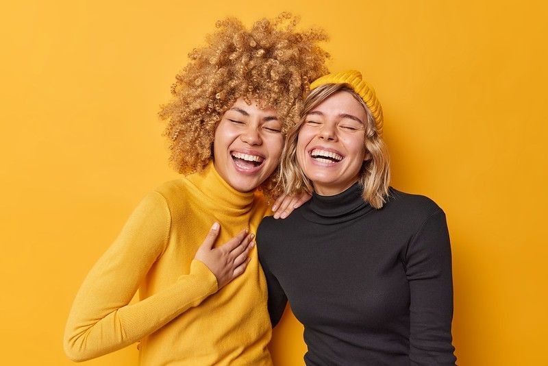 Young women laughing together dressed casually isolated over yellow wall