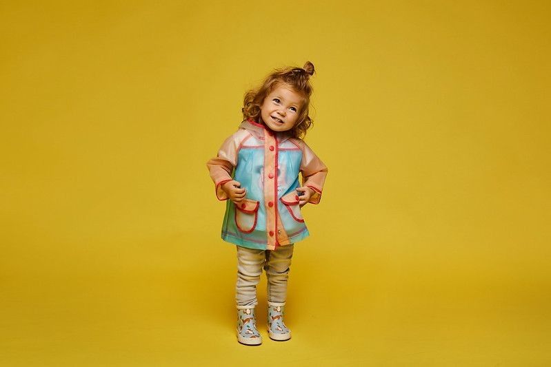 Cute little girl wearing raincoat and standing against yellow background