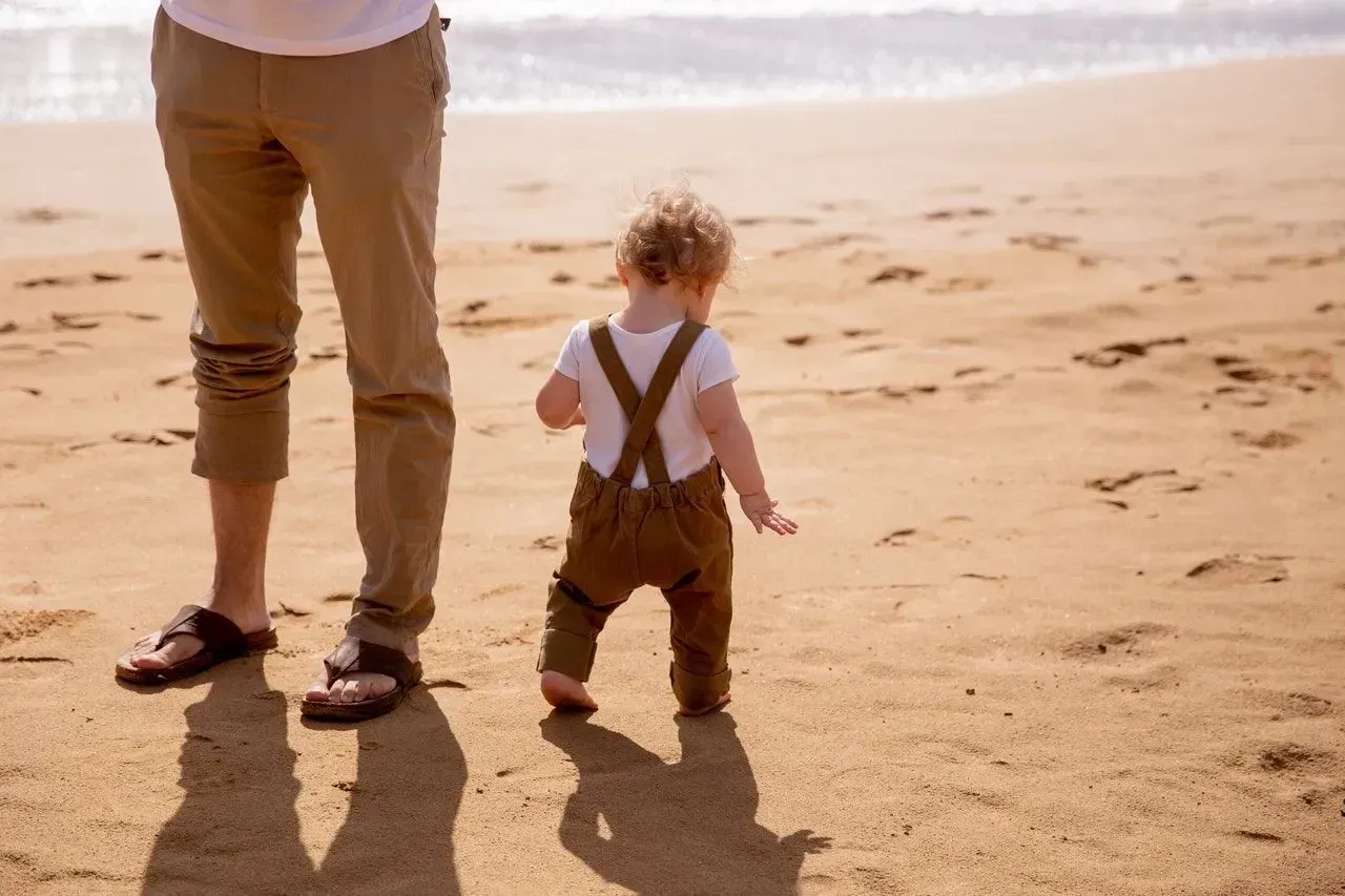 Little boy walking on beach sand in brown and white clothes with his father