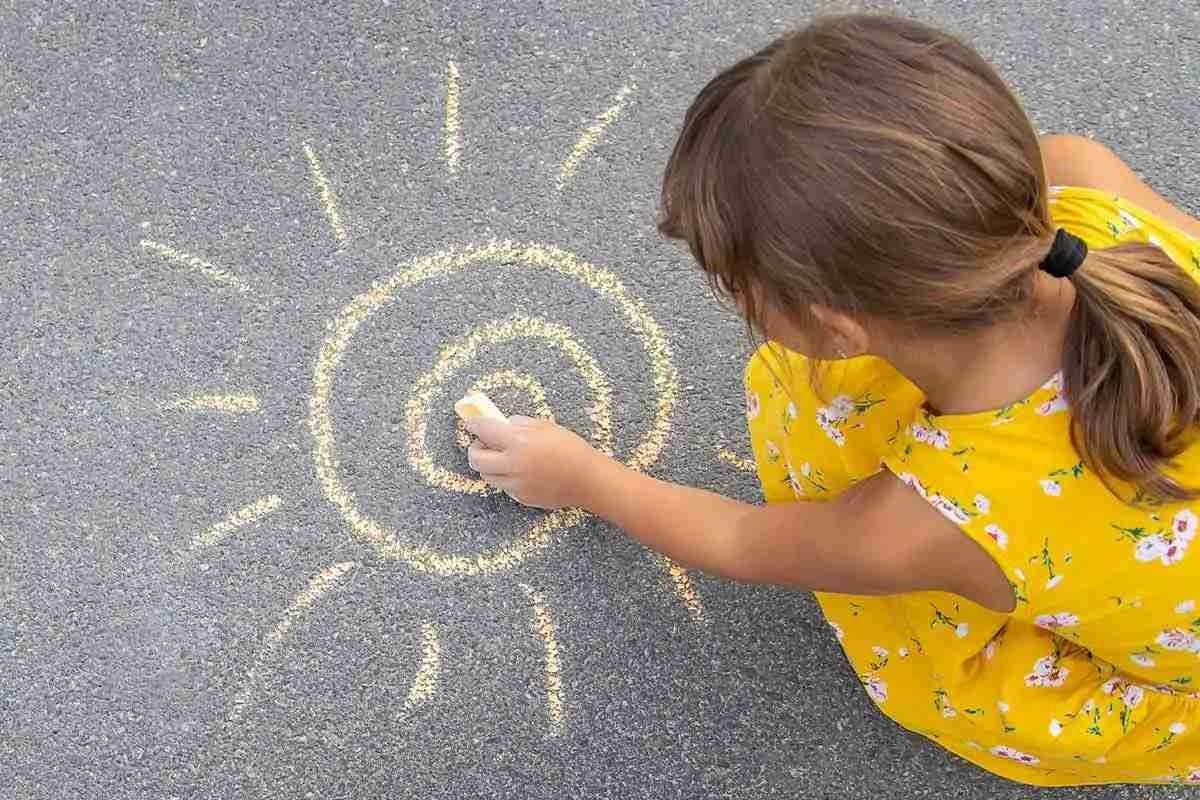 Little girl, wearing a yellow dress, crouching down drawing a sun, in yellow chalk, on the ground.