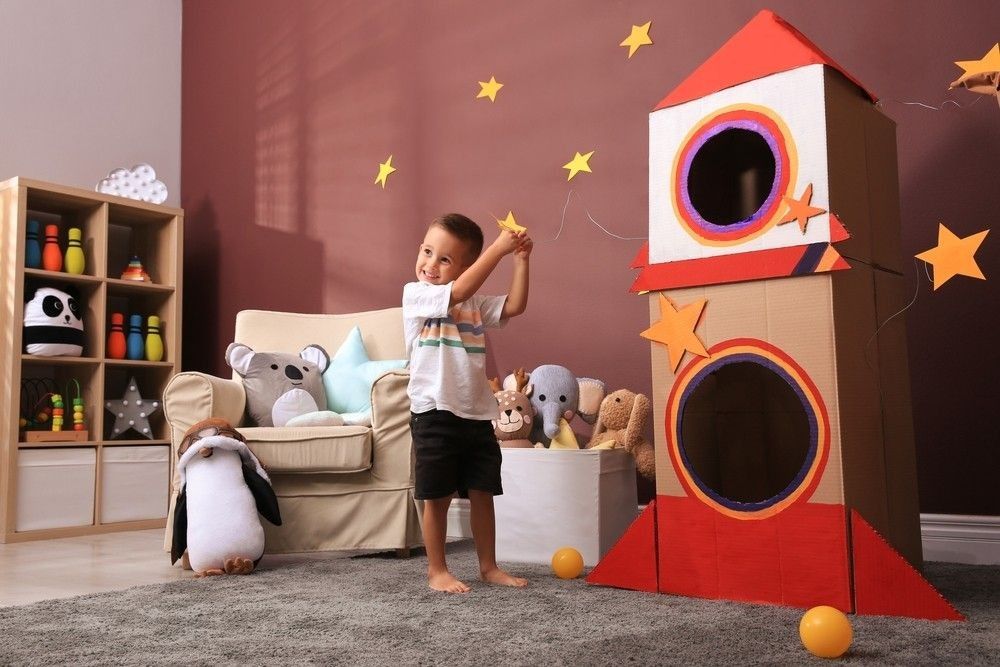 Boy playing on floor near cardboard rocket at home
