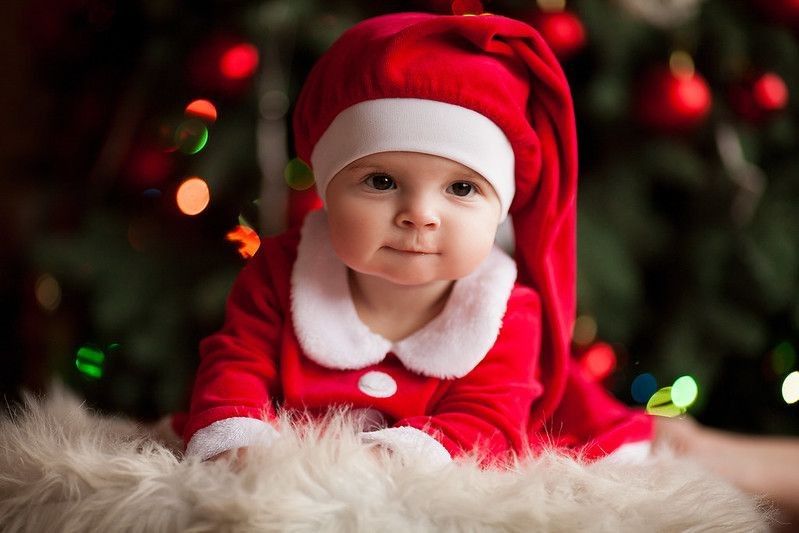 Cheerful baby girl in Santa Claus costume and hat lies on fur rug against background of Christmas decorations and lights.