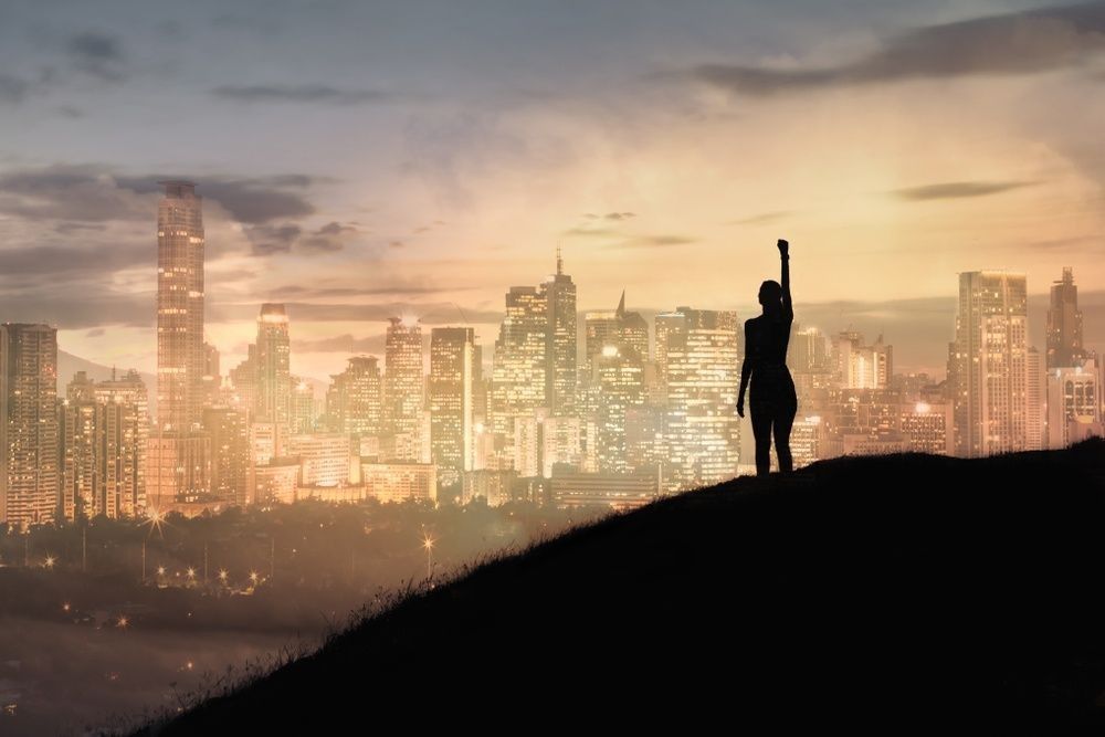 Strong woman with fist in the air standing on top a mountain overlooking the city.