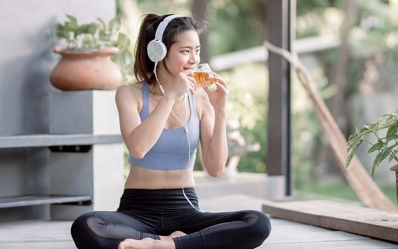 Asian beautiful and sportive woman drinking hot tea and wearing headphone while doing meditation in garden at morning time.